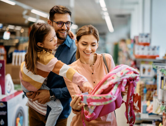 Little girl and her parents choosing backpack for school while shopping in store together.