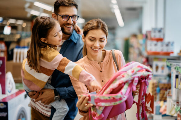 Little girl and her parents choosing backpack for school while shopping in store together.