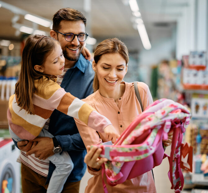 Little girl and her parents choosing backpack for school while shopping in store together.